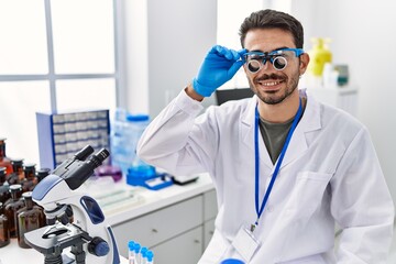 Wall Mural - Young hispanic man working at scientist laboratory wearing magnifying glasses looking positive and happy standing and smiling with a confident smile showing teeth