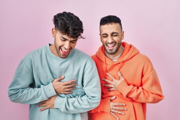 Wall Mural - Young hispanic gay couple standing over pink background smiling and laughing hard out loud because funny crazy joke with hands on body.