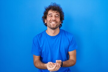 Canvas Print - Hispanic young man standing over blue background with hands together and crossed fingers smiling relaxed and cheerful. success and optimistic