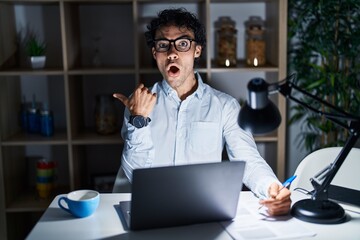 Canvas Print - Hispanic man working at the office at night surprised pointing with finger to the side, open mouth amazed expression.