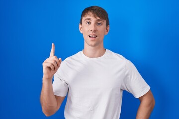 Canvas Print - Caucasian blond man standing over blue background pointing finger up with successful idea. exited and happy. number one.