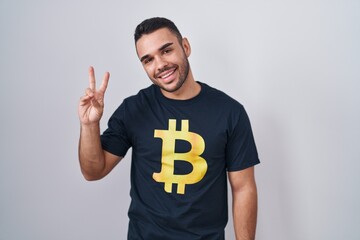 Poster - Young hispanic man wearing bitcoin t shirt smiling looking to the camera showing fingers doing victory sign. number two.