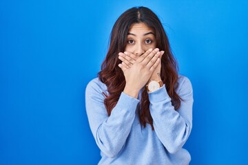 Sticker - Hispanic young woman standing over blue background shocked covering mouth with hands for mistake. secret concept.