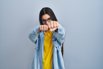 Canvas Print - Young hispanic woman standing over blue background punching fist to fight, aggressive and angry attack, threat and violence