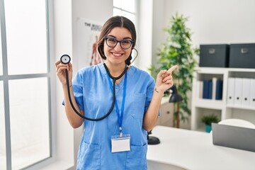 Canvas Print - Young hispanic doctor woman wearing stethoscope at the clinic smiling happy pointing with hand and finger to the side