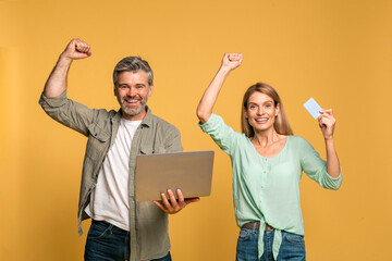 Wall Mural - Excited caucasian couple with credit card and laptop making gesture of victory and success, enjoying sales