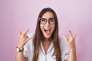 Poster - Young brunette woman wearing glasses standing over pink background shouting with crazy expression doing rock symbol with hands up. music star. heavy concept.