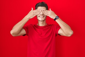 Sticker - Young hispanic man standing over red background covering eyes with hands smiling cheerful and funny. blind concept.