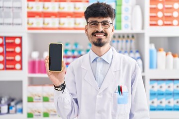 Sticker - Young hispanic man with beard working at pharmacy drugstore showing smartphone screen looking positive and happy standing and smiling with a confident smile showing teeth