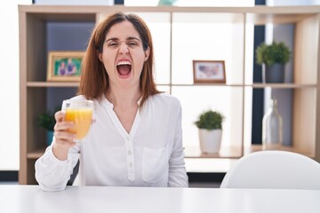 Wall Mural - Brunette woman drinking glass of orange juice angry and mad screaming frustrated and furious, shouting with anger. rage and aggressive concept.