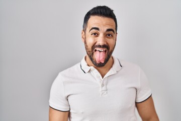 Poster - Young hispanic man with beard wearing casual clothes over white background sticking tongue out happy with funny expression. emotion concept.