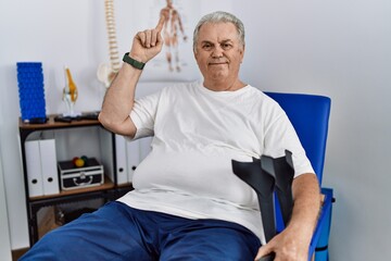 Wall Mural - Senior caucasian man at physiotherapy clinic holding crutches smiling amazed and surprised and pointing up with fingers and raised arms.