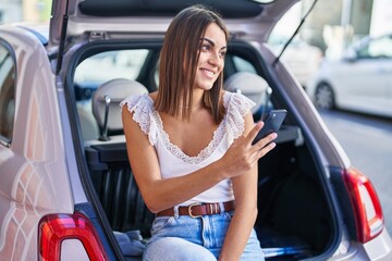 Young beautiful hispanic woman using smartphone sitting on car trunk at street