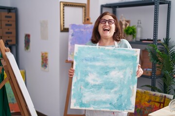 Poster - Senior woman with glasses holding canvas at art studio smiling and laughing hard out loud because funny crazy joke.