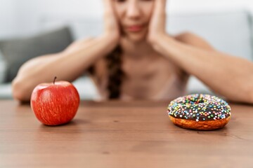 Wall Mural - Young caucasian woman choosing healthy or unhealthy food sitting on sofa at home