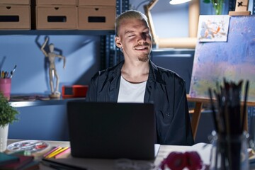 Sticker - Young caucasian man using laptop at night at art studio winking looking at the camera with sexy expression, cheerful and happy face.