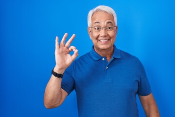 Poster - Middle age man with grey hair standing over blue background smiling positive doing ok sign with hand and fingers. successful expression.