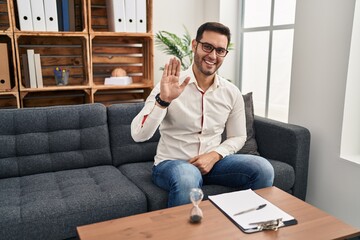 Canvas Print - Young hispanic man with beard working at consultation office waiving saying hello happy and smiling, friendly welcome gesture