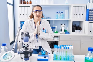 young hispanic woman working at scientist laboratory begging and praying with hands together with ho