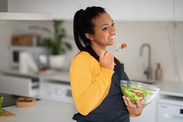 Cheerful pretty millennial african american lady in apron eat fresh vegetable salad in white kitchen interior