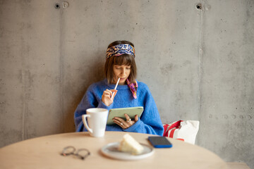 Wall Mural - Young stylish woman works on a digital tablet while sitting on concrete wall background at modern coffee shop. Concept of remote creative work online