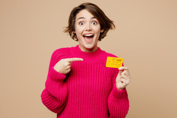 Wall Mural - Young happy cheerful amazed caucasian woman wear pink sweater hold in hand point index finger on mock up of credit bank card isolated on plain pastel light beige background. People lifestyle concept.