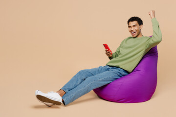 Wall Mural - Full body satisfied young man of African American ethnicity wear green sweatshirt sit in bag chair hold in hand use mobile cell phone do winner gesture isolated on plain pastel light beige background.