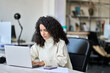 © insta_photos - Young latin professional business woman worker sitting at desk working on marketing data online on laptop in modern corporate office. Female worker using computer technology typing browsing web.