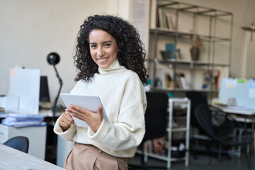 Smiling latin young professional business woman corporate marketing manager, female worker holding digital tablet computer fintech tab at work standing in modern company office looking at camera.