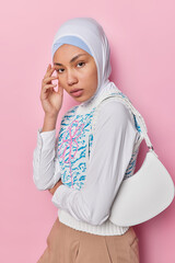 Wall Mural - Studio shot of charming beautiful Arabic woman touches face gently has healthy skin wears hijab white shirt and vest poses with bag on shoulder isolated over pink background. Religious female model