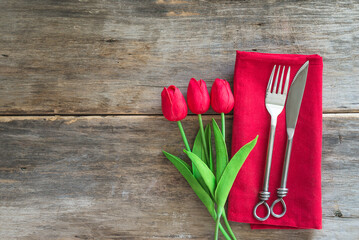 Festive place setting for romantic dinner; forged fork and knife on red napkin, bouquet of red tulips on old wooden background; space for text