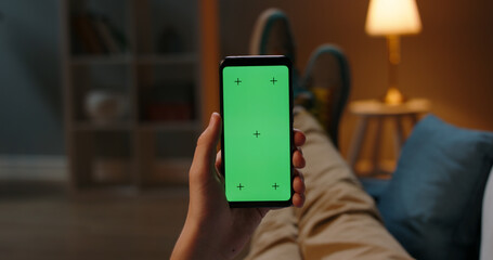 Man lying on couch using smartphone with chroma key green screen at night, scrolling through social media or online shop - internet, communications concept close up 