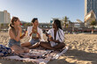 © PintoArt - group of female friends enjoying a day at the beach, in summer, multiracial female friends eat refreshing fruit on the sand on the beach on a sunny day