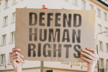 The Phrase " Defend Human Rights " Is On A Banner In Men's Hands With Blurred Background. Secure. Stand. Support. Champion. Fight. Help. Rally. Shield. Strengthen. Verify. Watch. Brave. Endure. Trust