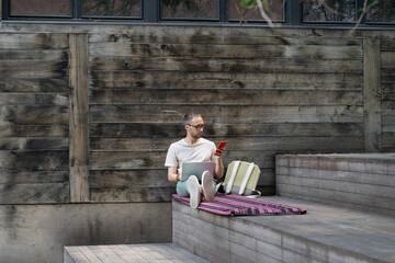 Young enterprising man programmer sits outdoors with laptop and phone creating software to sell online. Successful guy IT engineer located near wooden wall of building on street in summer weather