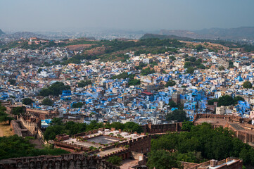 Wall Mural - Top view of Mehrangarh fort with distant view of blue city Jodhpur, Rajasthan, India. Blue sky with white clouds in the background. One of the seven gates to enter the huge fort is seen below,