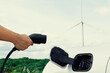 Progressive natural scenic with windmill generator where hand insert charging plug to electric vehicle from charging station with natural background. EV car powered by wind turbine electric generator.