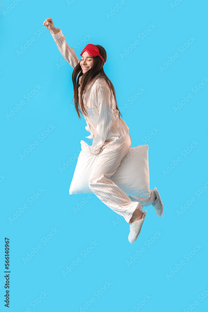 Young woman with sleeping mask and soft pillow jumping on blue background