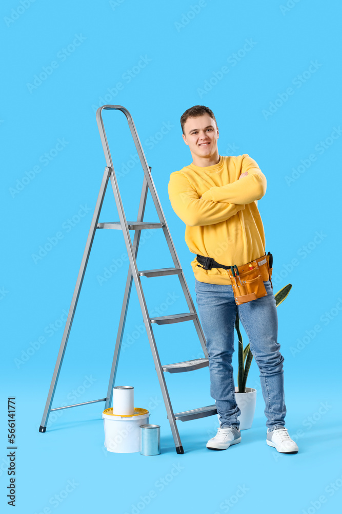 Young man with ladder and cans of paint on blue background