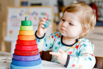 Cute beautiful little baby girl playing with educational toys at home or nursery, indoors. Happy healthy child having fun with colorful wooden rainbow toy pyramid. Kid learning different skills.