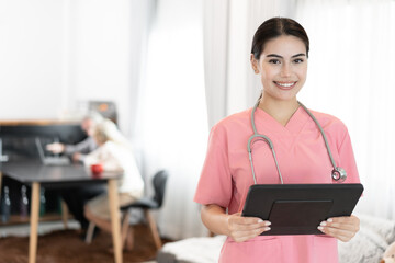 Wall Mural - Beautiful nurse holding tablet computer with older caucasian woman and man background at home