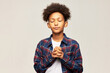 Studio image of cute african american teen boy kid praying for winning school competition or making wish, standing against gray studio background with interlocked fingers and closed eyes