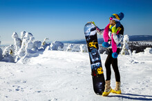 Girl Snowboarder Enjoys The Ski Resort. Beautiful Woman In Winter Outfit Is Posing With Snowboard In The Mountains. Winter Sports. Snowboarding.