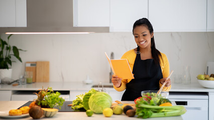 Wall Mural - Smiling pretty millennial black female in apron making salad, reading new recipe in notebook book