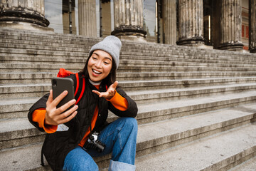 Wall Mural - Smiling asian woman making video call via smartphone and gesturing while sitting on stairs