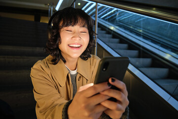 Wall Mural - Close up portrait of smiling asian girl student, listens music in headphones and looks at mobile phone, uses smartphone, sits on staircase in public place