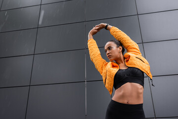 Wall Mural - low angle view of african american sportswoman in sports bra and yellow puffer jacket warming up outside.