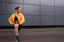 Full Length Of Stylish African American Sportswoman In Bike Shorts And Yellow Puffer Jacket Working Out Near Grey Building.