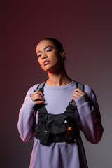 Wall Mural - young african american woman in sweatshirt and vest looking at camera on grey and purple.