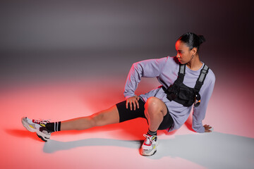 Wall Mural - full length of african american woman in bike shorts and sweatshirt posing on grey background with red light.